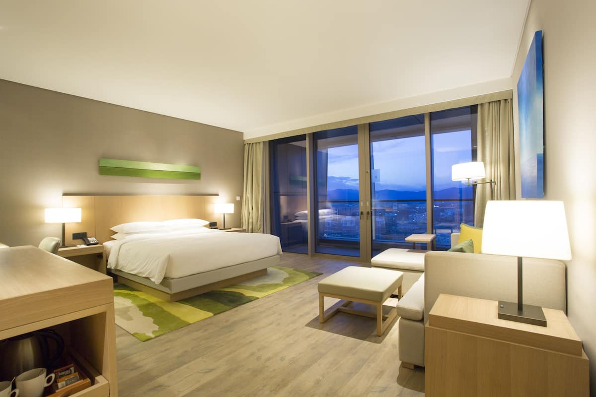 Een moderne hotelkamer met een groot bed, een bureau, een stoel, een tafeltje en uitzicht op de stad door grote ramen.