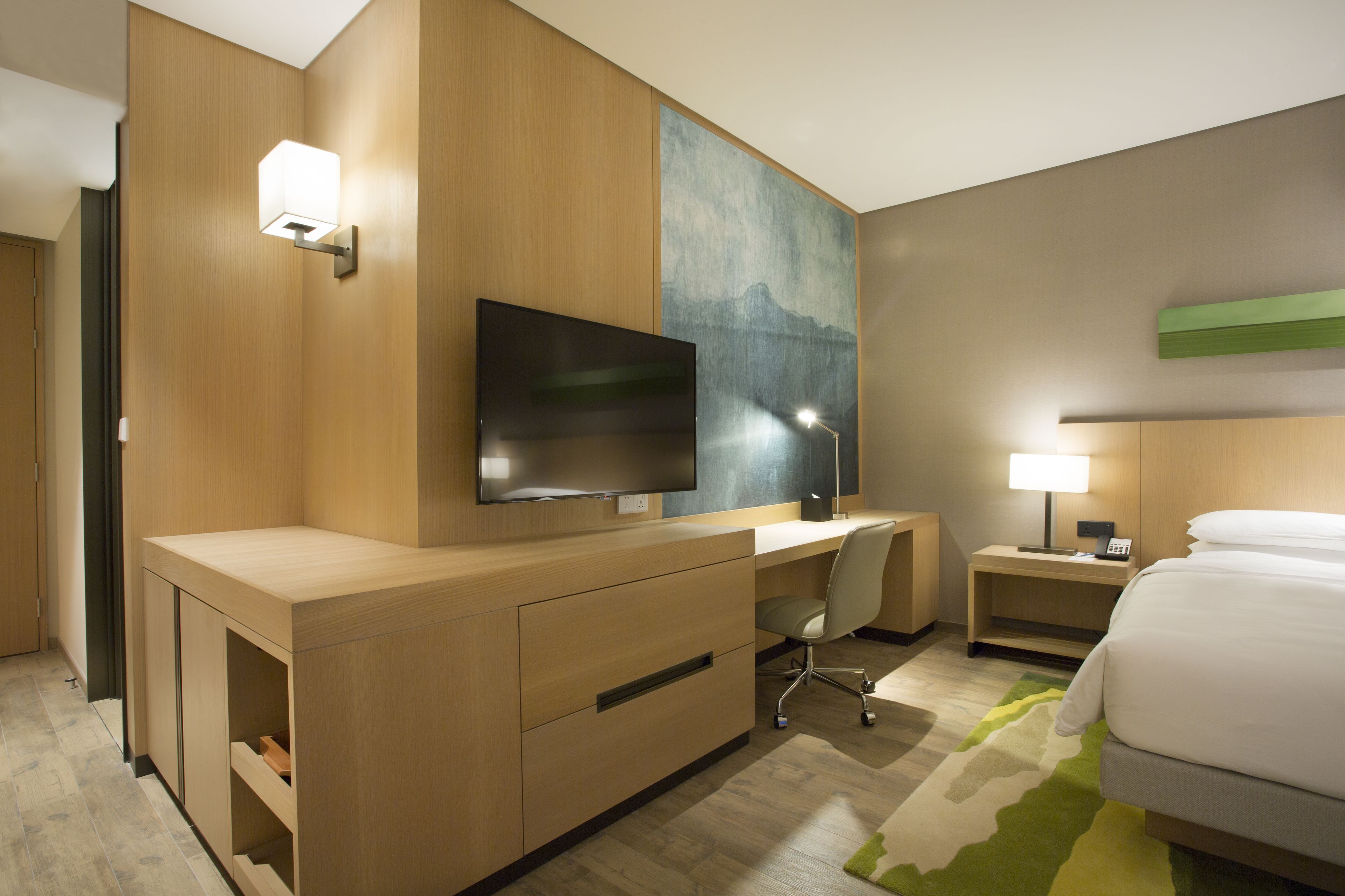 A hotel room with a wooden desk, a flat-screen TV, a bed, a nightstand with a lamp, and a painting on the wall.