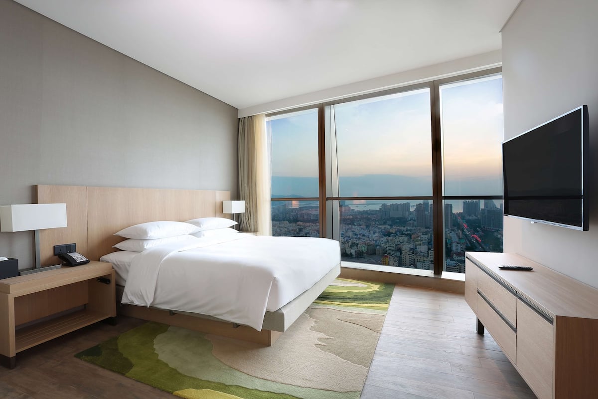 Een moderne hotelkamer met een groot bed, een nachtkastje met een lamp, een aan de muur bevestigde televisie en uitzicht op de stad door grote ramen.