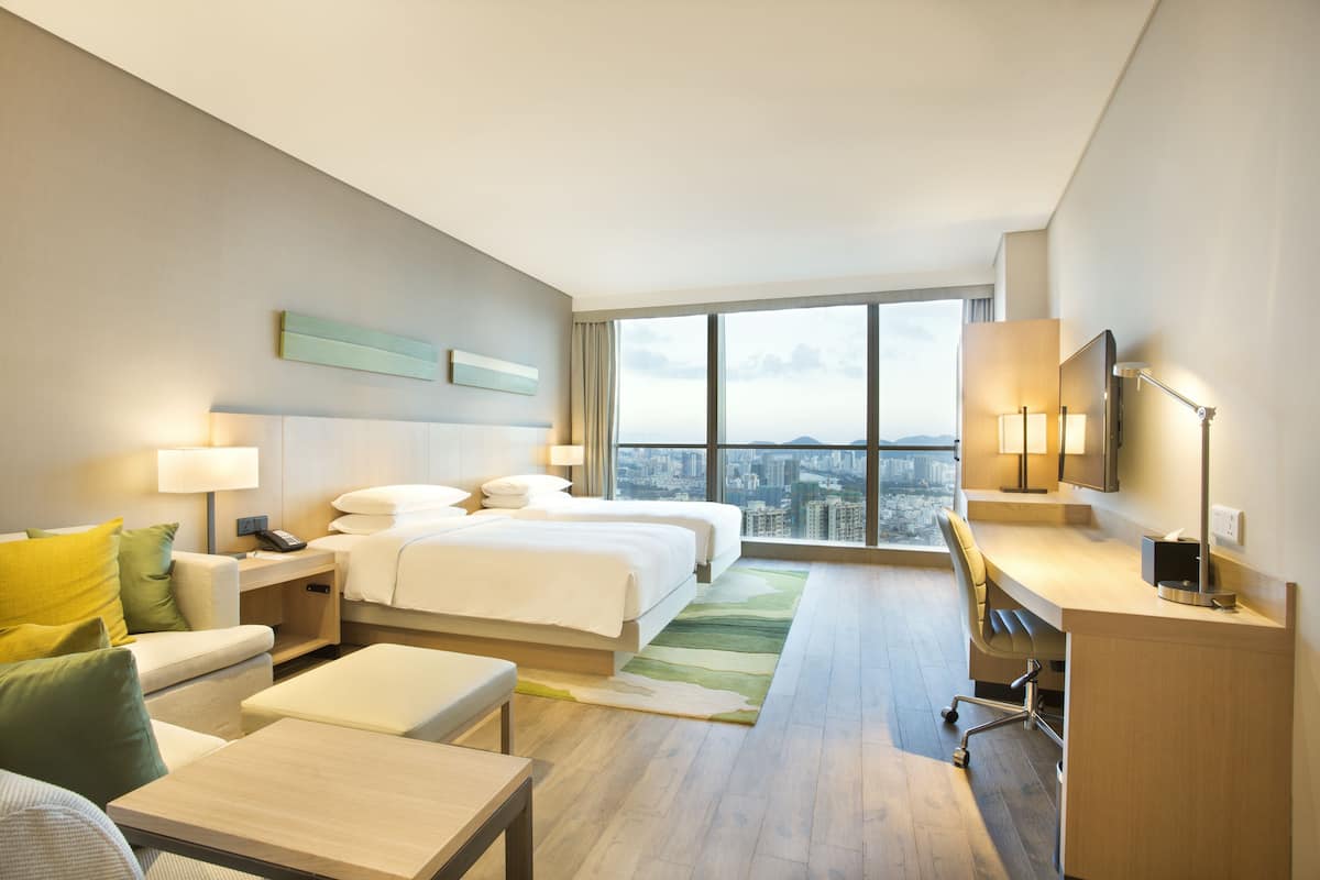 Hotelkamer met twee bedden, een bureau, een stoel, een bank en een groot raam met uitzicht over de stad.