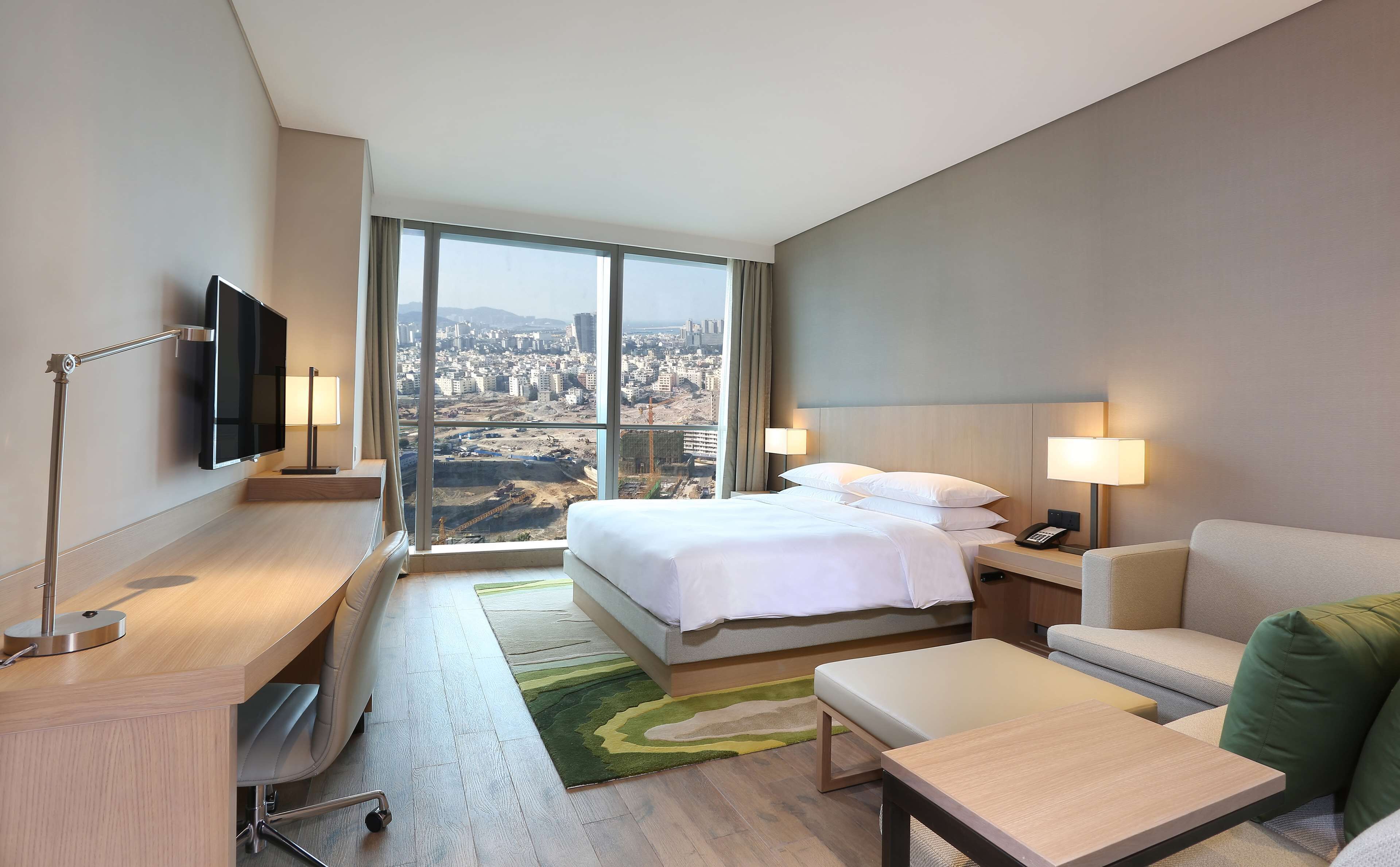 A hotel room with a large bed, a desk with a chair, a TV mounted on the wall, and a view of a cityscape through large windows.