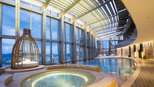 Une piscine intérieure moderne au design incurvé, dotée de grandes fenêtres offrant une vue sur la ville et d’un espace central avec un jacuzzi.