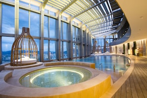 Een modern binnenzwembad met een golvend ontwerp, grote ramen met uitzicht op de stad, en een centraal gelegen jacuzzi.
