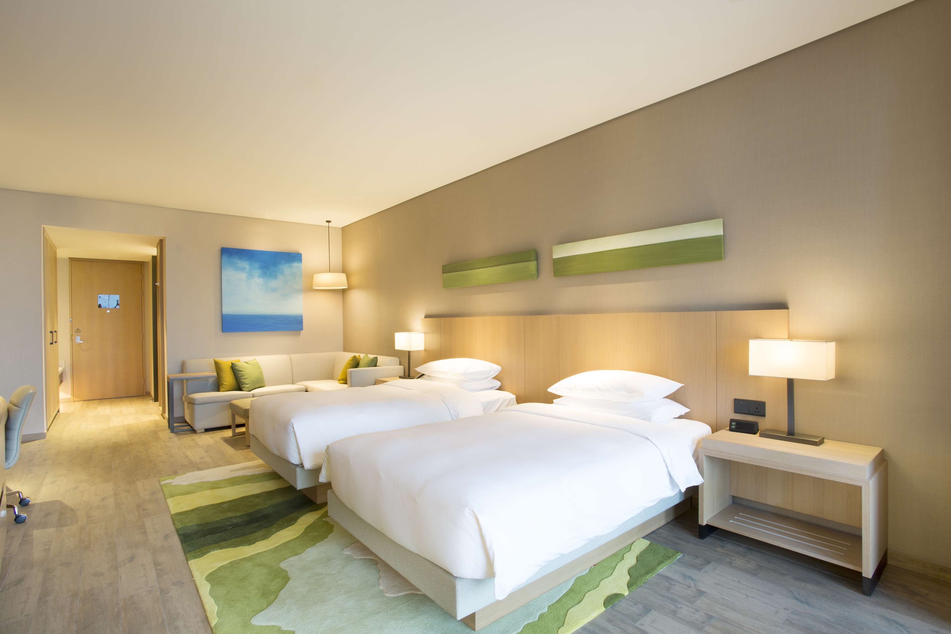 A hotel room with two beds, a desk, a chair, a nightstand with a lamp, and a painting on the wall.