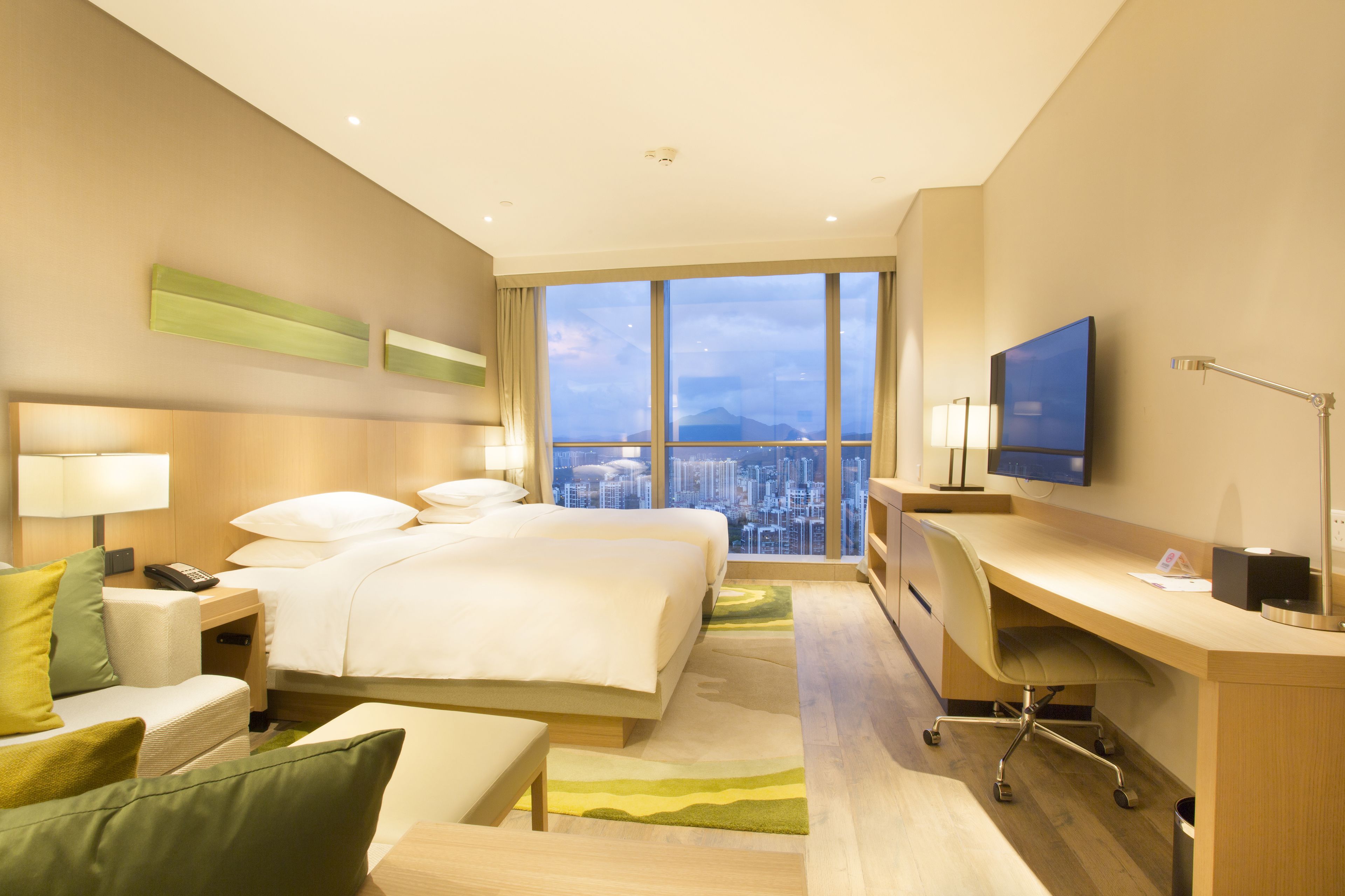 A hotel room with a large bed, a desk, a TV, and a view of a cityscape.