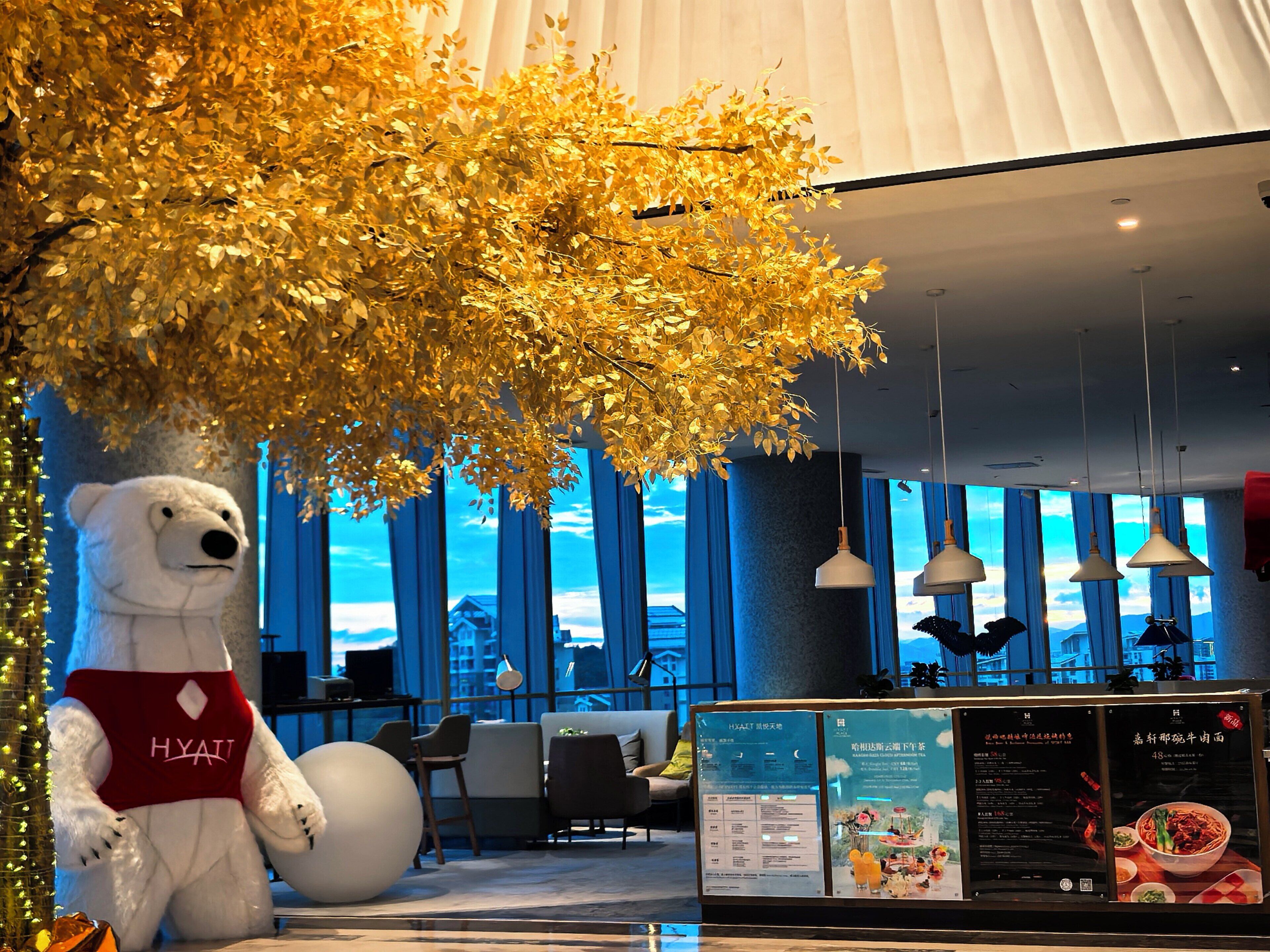 Ein flauschiges, weißes Maskottchen in Form eines Bären mit roter Schürze in einer modernen Hotelhalle mit großen Fenstern, einer Sitzecke und einer Speisekarte.