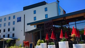 A modern hotel with a white facade, multiple windows, and a covered outdoor seating area with red umbrellas. - ibis budget Annecy Poisy (Poisy)