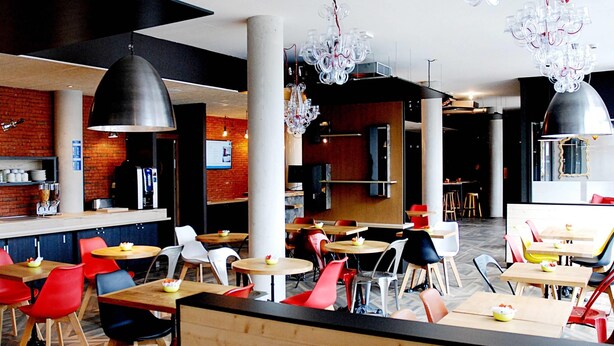 A modern restaurant interior with high ceilings, exposed brick walls, and a mix of wooden and metal furniture. - ibis budget Annecy Poisy (Poisy)