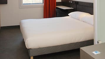 Ein Hotelzimmer mit Bett, Schreibtisch, einem Fenster mit Vorhängen und einer Wandsteckdose.