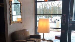 A room with a brown leather sofa, a large floor lamp, a small table with books, a mirror, and a view of parked cars outside. - ibis budget Annecy Poisy (Poisy)