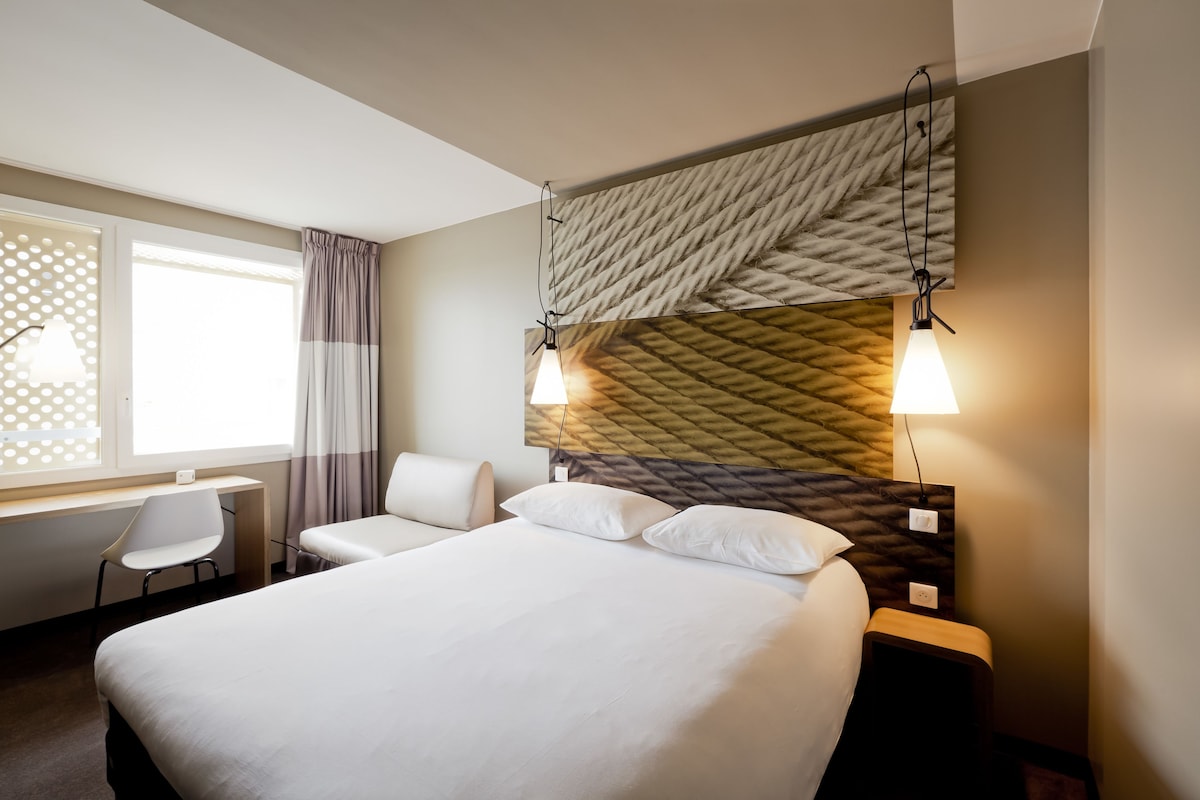 Habitación de hotel con una cama grande, un escritorio y una silla. Se aprecia una ventana con cortinas, y una pared con textura decorativa.