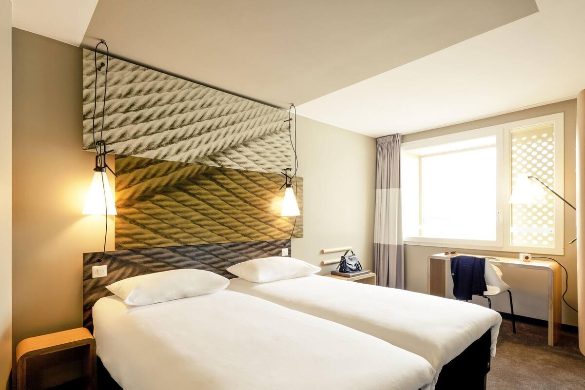 Une chambre d’hôtel avec deux lits, un bureau, une chaise et une fenêtre avec des rideaux.