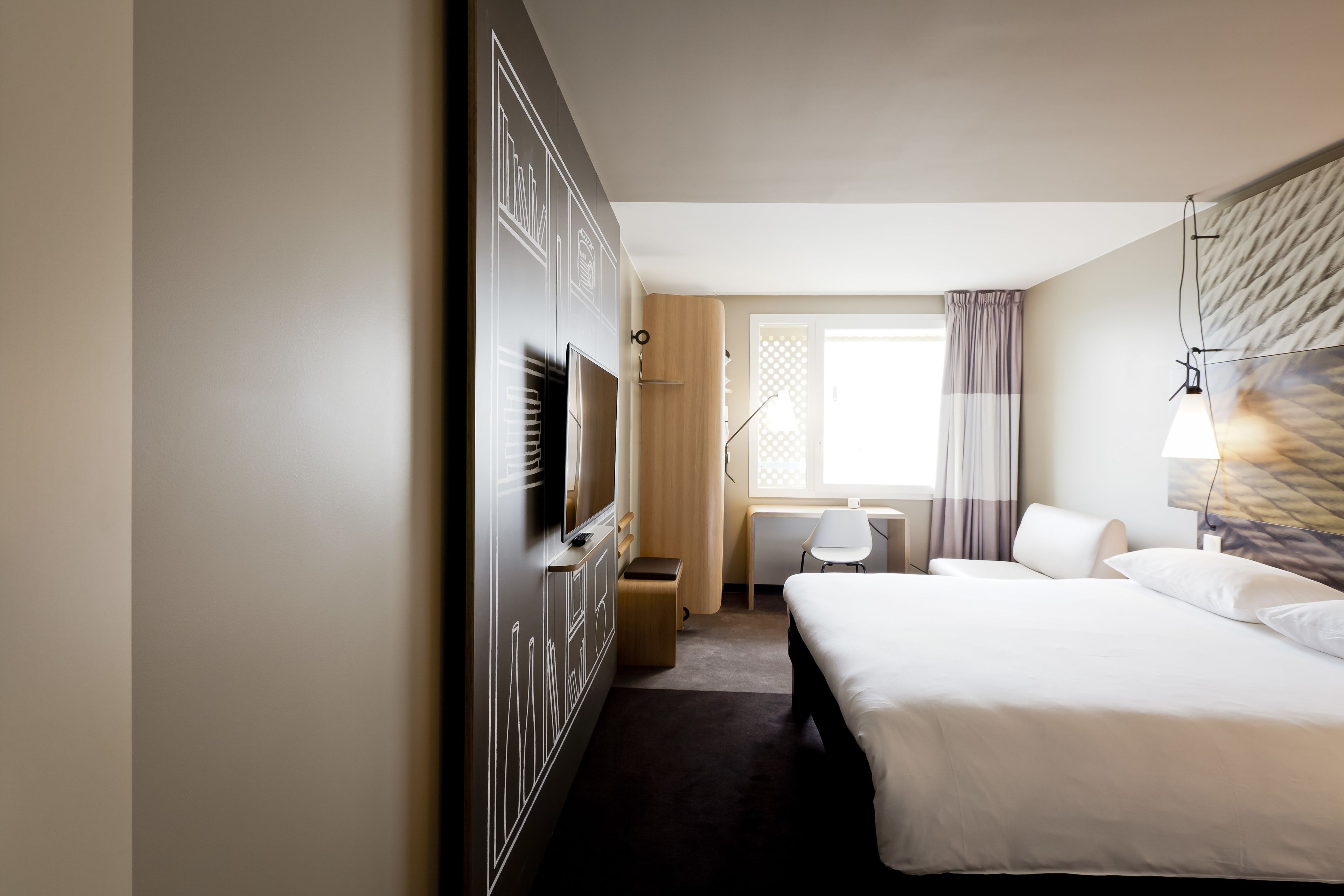 A hotel room with a large bed, a desk, a chair, a TV, and a window with curtains.