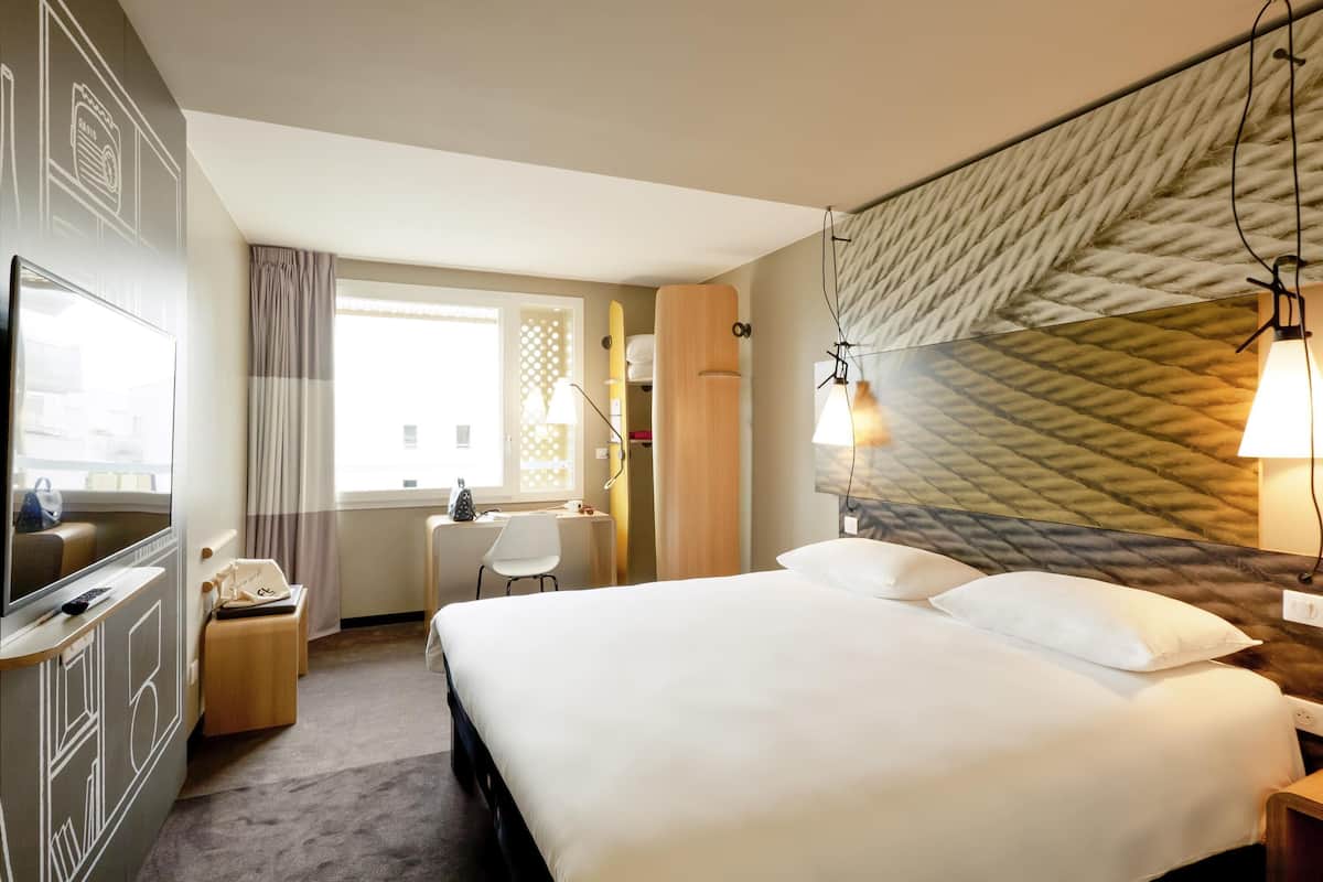 Une chambre d’hôtel avec un lit, un bureau et une chaise, une télévision et une fenêtre avec des rideaux.