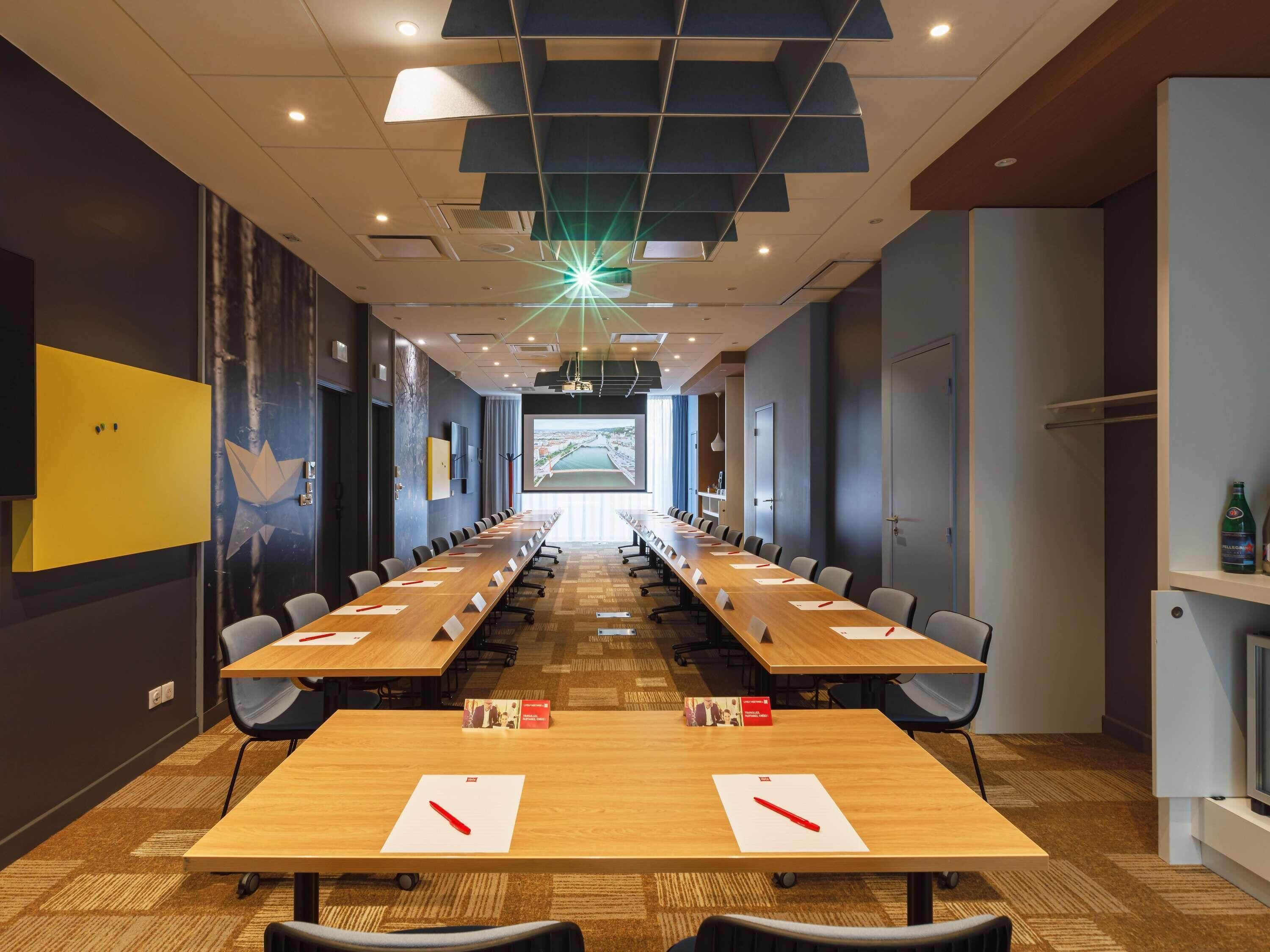 A conference room with long tables, chairs, a projector screen, and a large window.