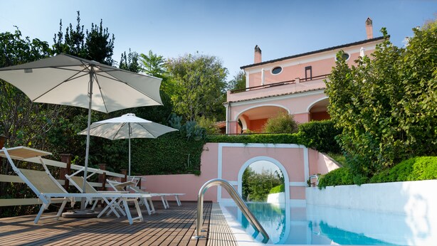 Un espace piscine avec des chaises longues et des parasols, une vue dégagée sur la piscine et un bâtiment à la façade rose.