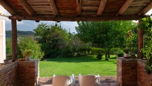 Une terrasse couverte avec deux chaises blanches, un mur de briques et une vue sur une pelouse verdoyante et des arbres.
