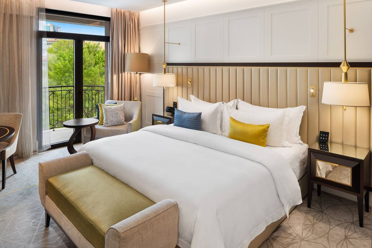 A hotel room with a large bed, a sitting area with a chair and a small table, a nightstand with a lamp, and a view of greenery through the window.