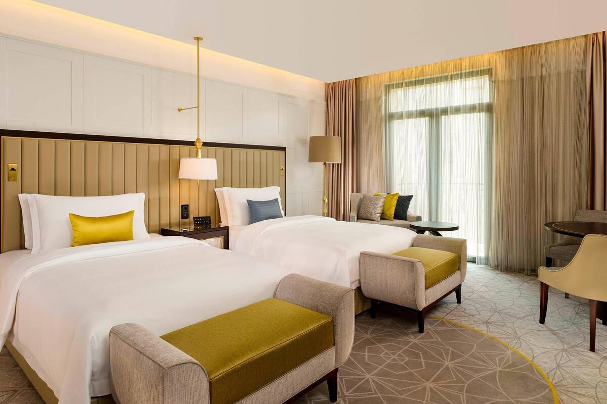 A hotel room with two beds, a sitting area with a table and chairs, and a large window with curtains.