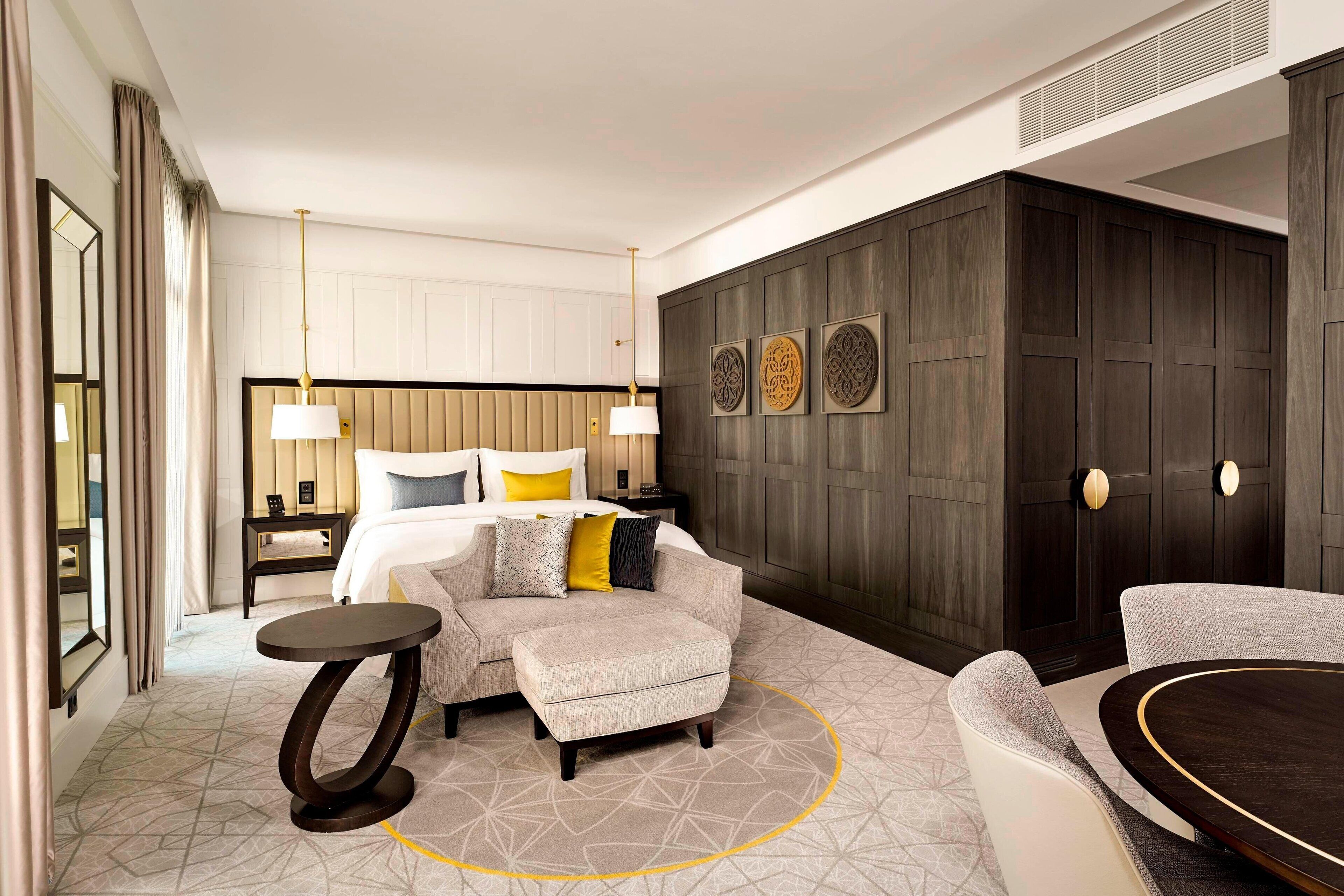 A hotel room with a bed, two armchairs, a small table, a decorative wall panel, and a ceiling vent.