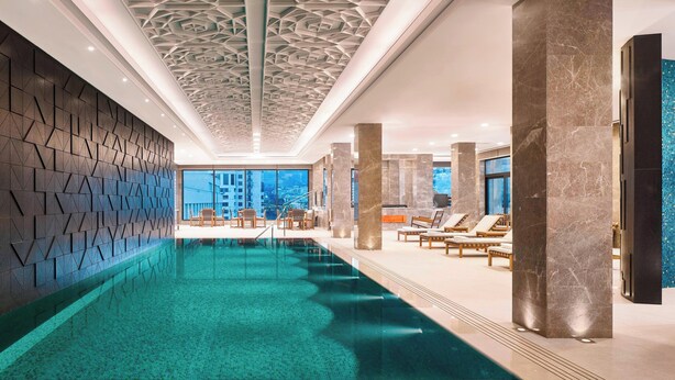 An indoor swimming pool with geometric ceiling design, marble columns, and lounge chairs.