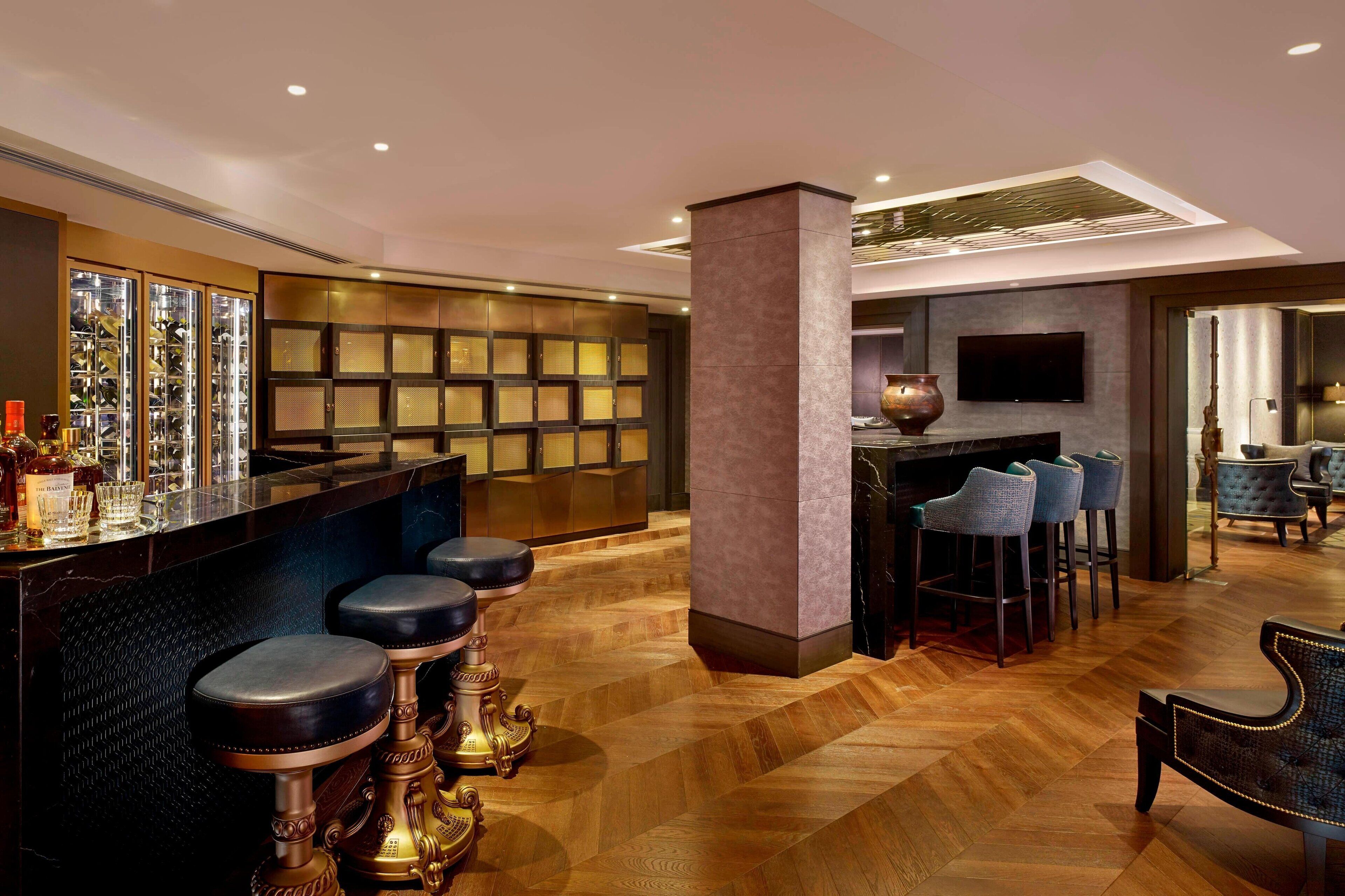 A modern bar area with a polished wooden floor, a bar counter with stools, and a television mounted on the wall.