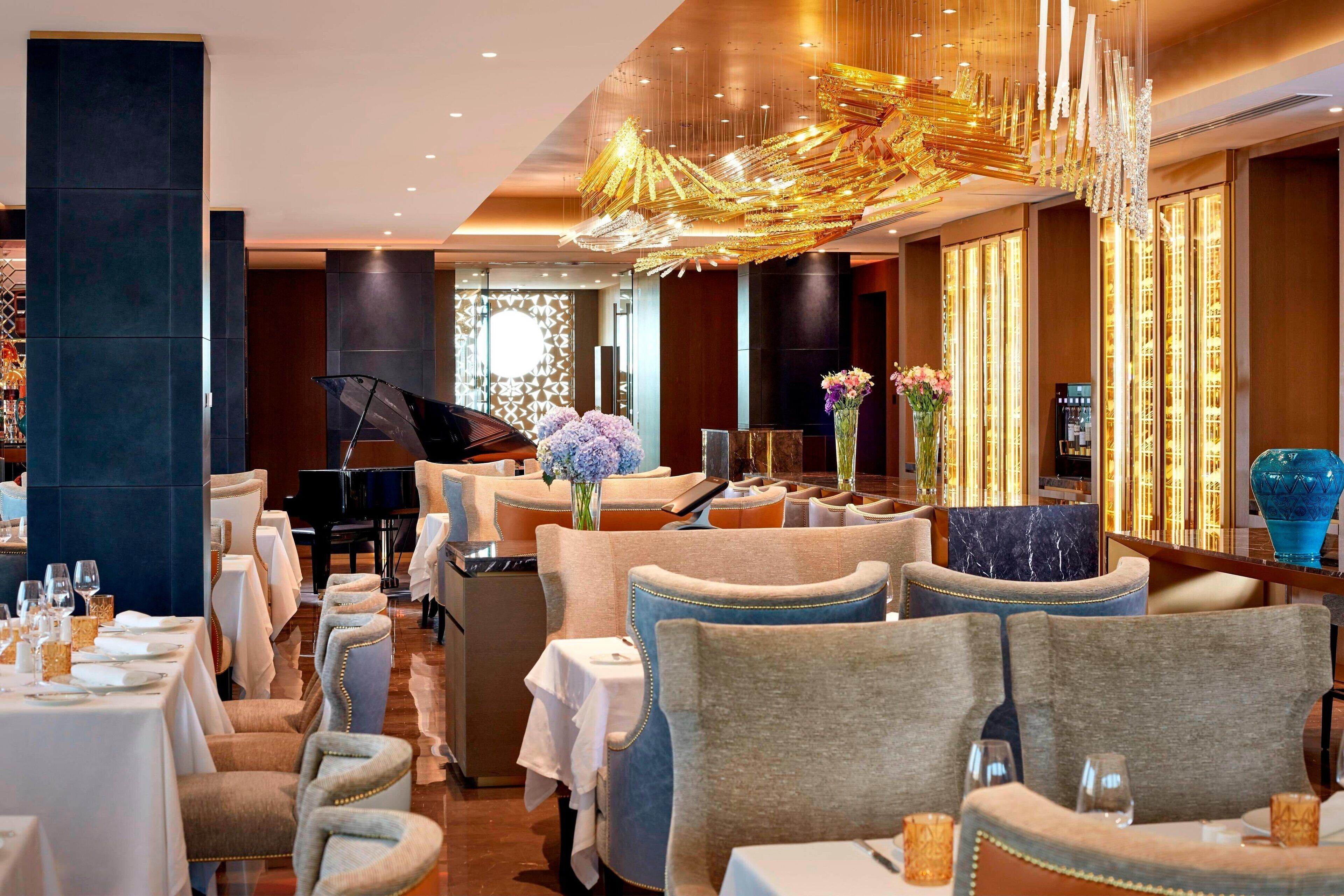 A restaurant with tables set for dining, chairs, a grand piano, and a decorative chandelier.