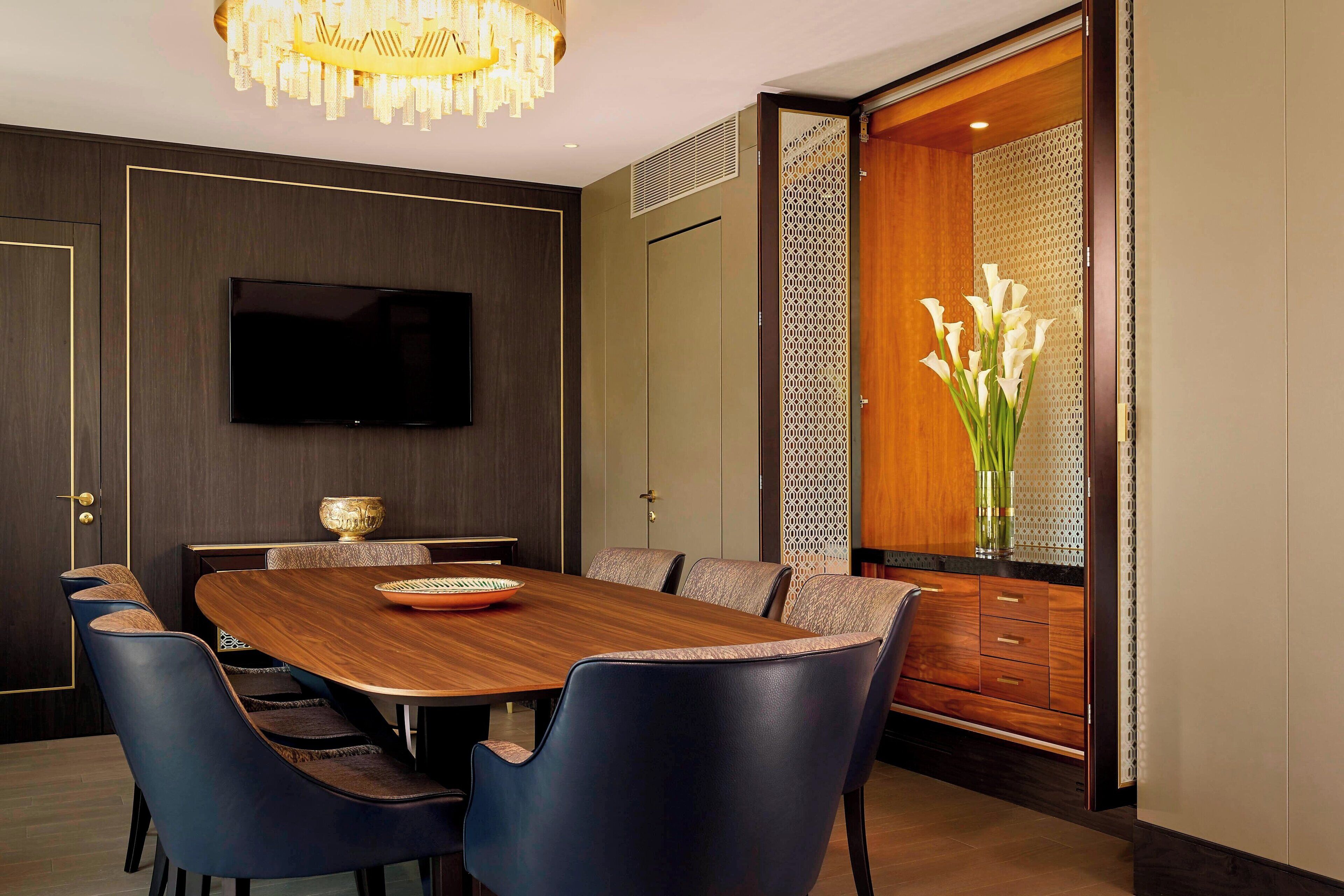 A modern dining area with a wooden table and blue chairs, a flat-screen TV on the wall, and a decorative vase with white flowers.