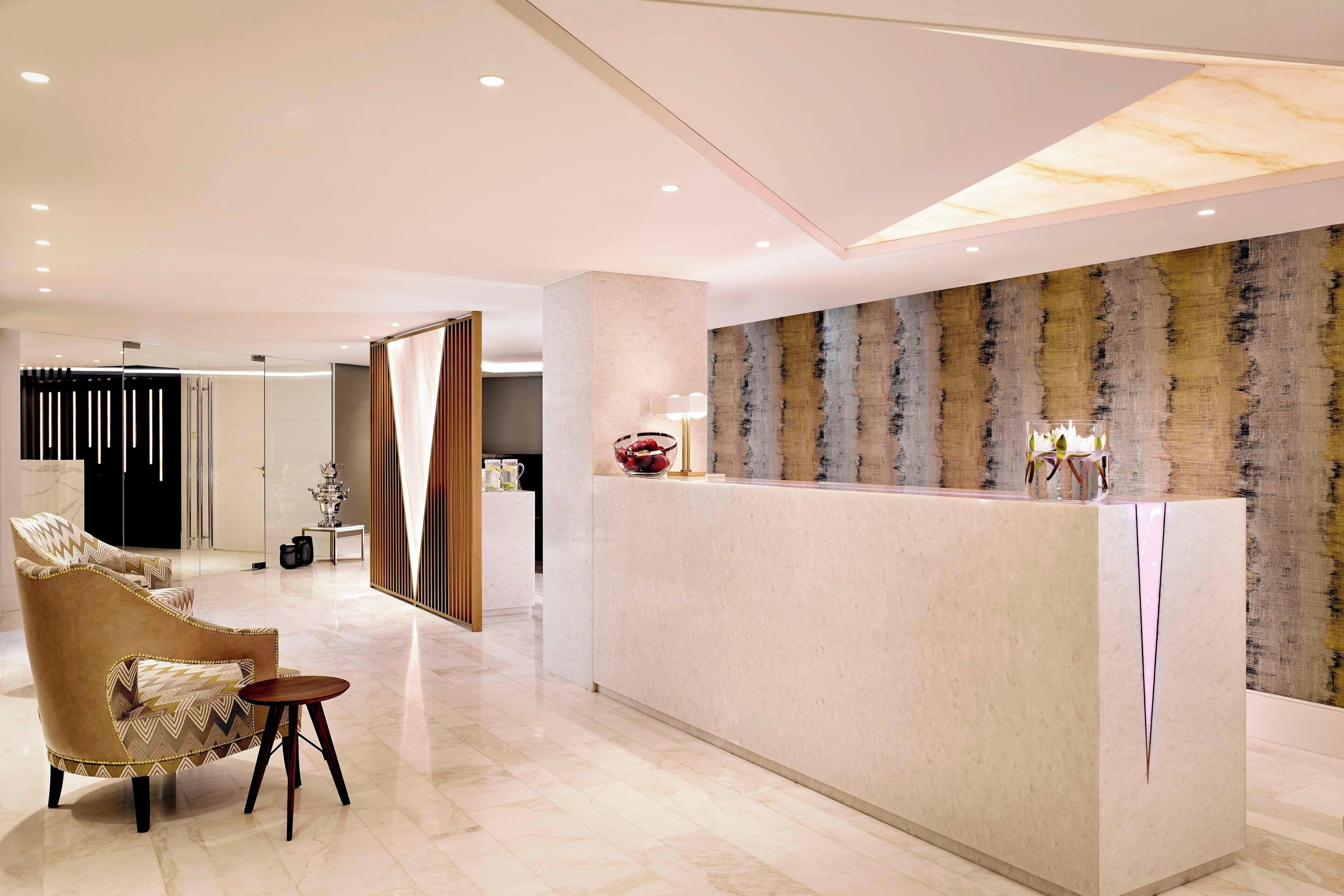 A modern hotel lobby with a reception desk, a seating area with a sofa and armchair, and a decorative wall with vertical patterns.