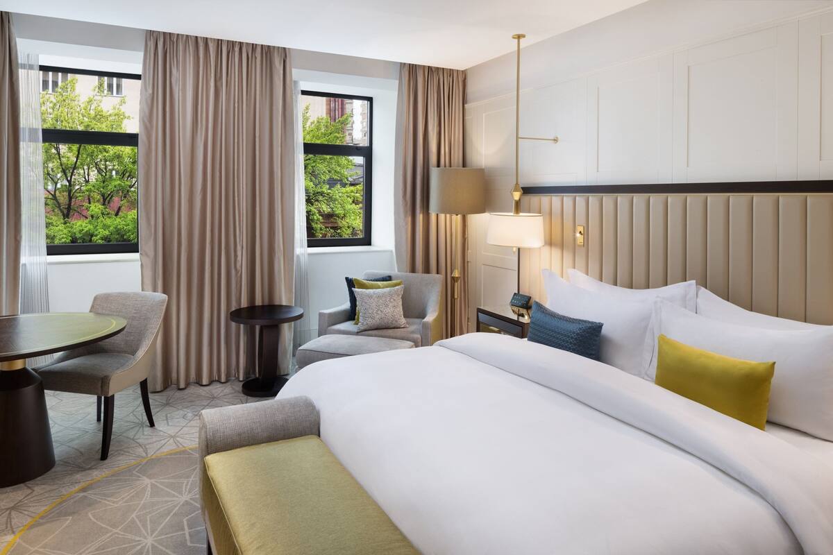 A hotel room with a bed, a chair, a small table, a lamp, and a view of greenery outside.