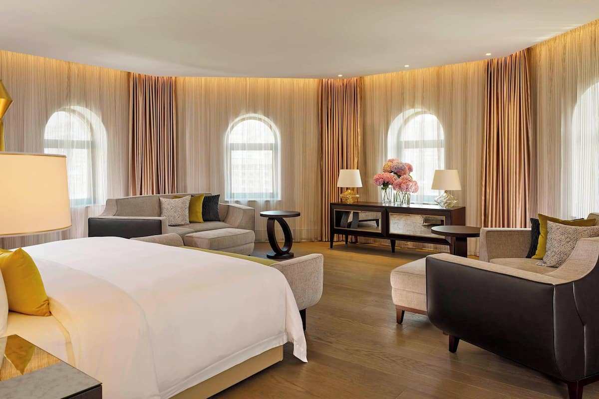 A hotel room with a large bed, two armchairs, a sofa, a small table, and a desk with lamps and flowers.