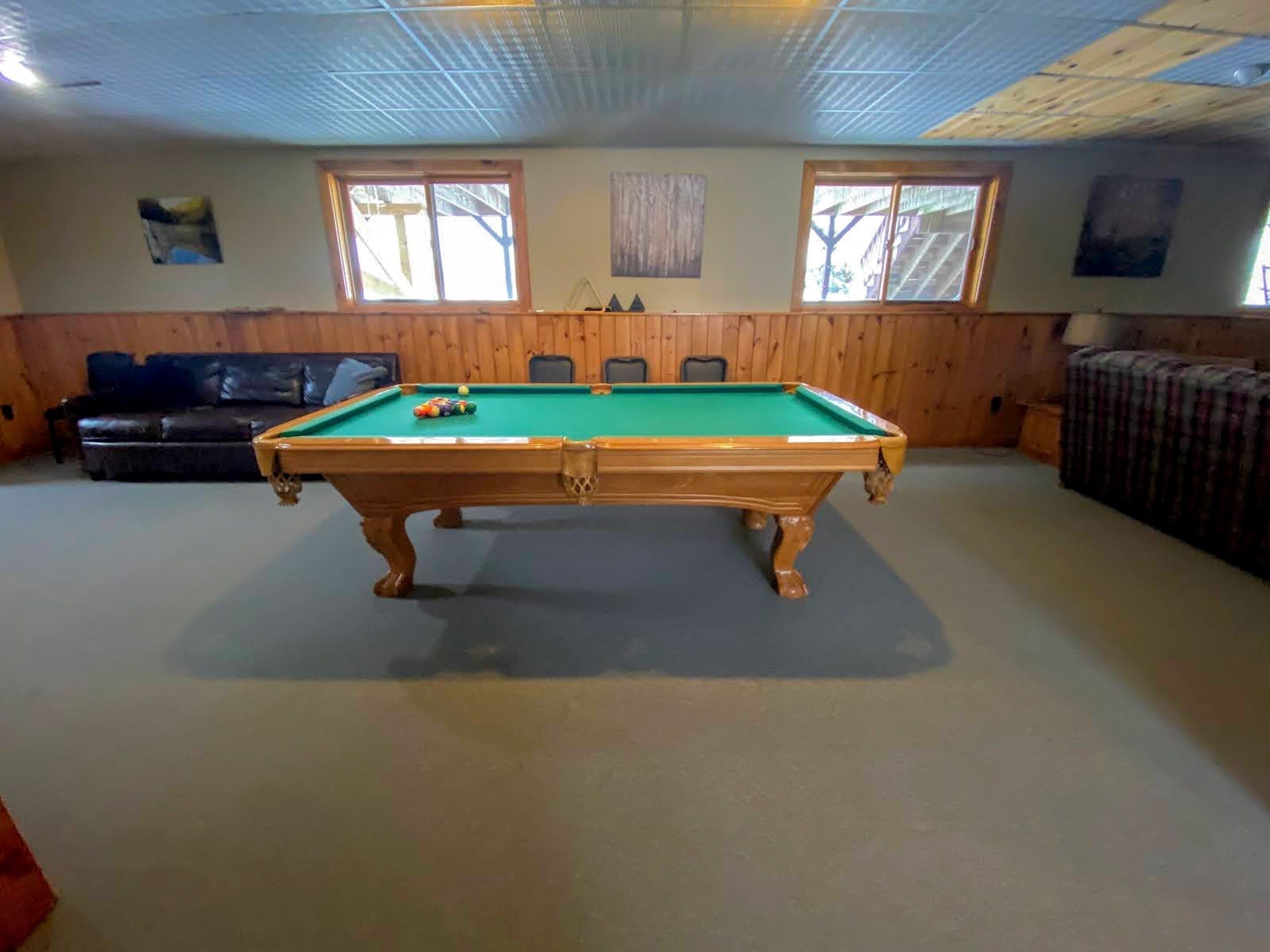 Une table de billard dans une pièce aux murs lambrissés, avec deux fenêtres et un canapé.