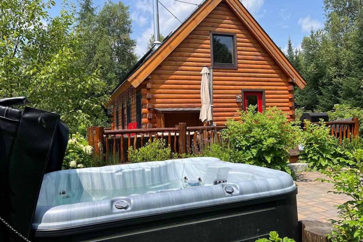 Une cabane en rondins avec un jacuzzi à l’avant, entourée de verdure.
