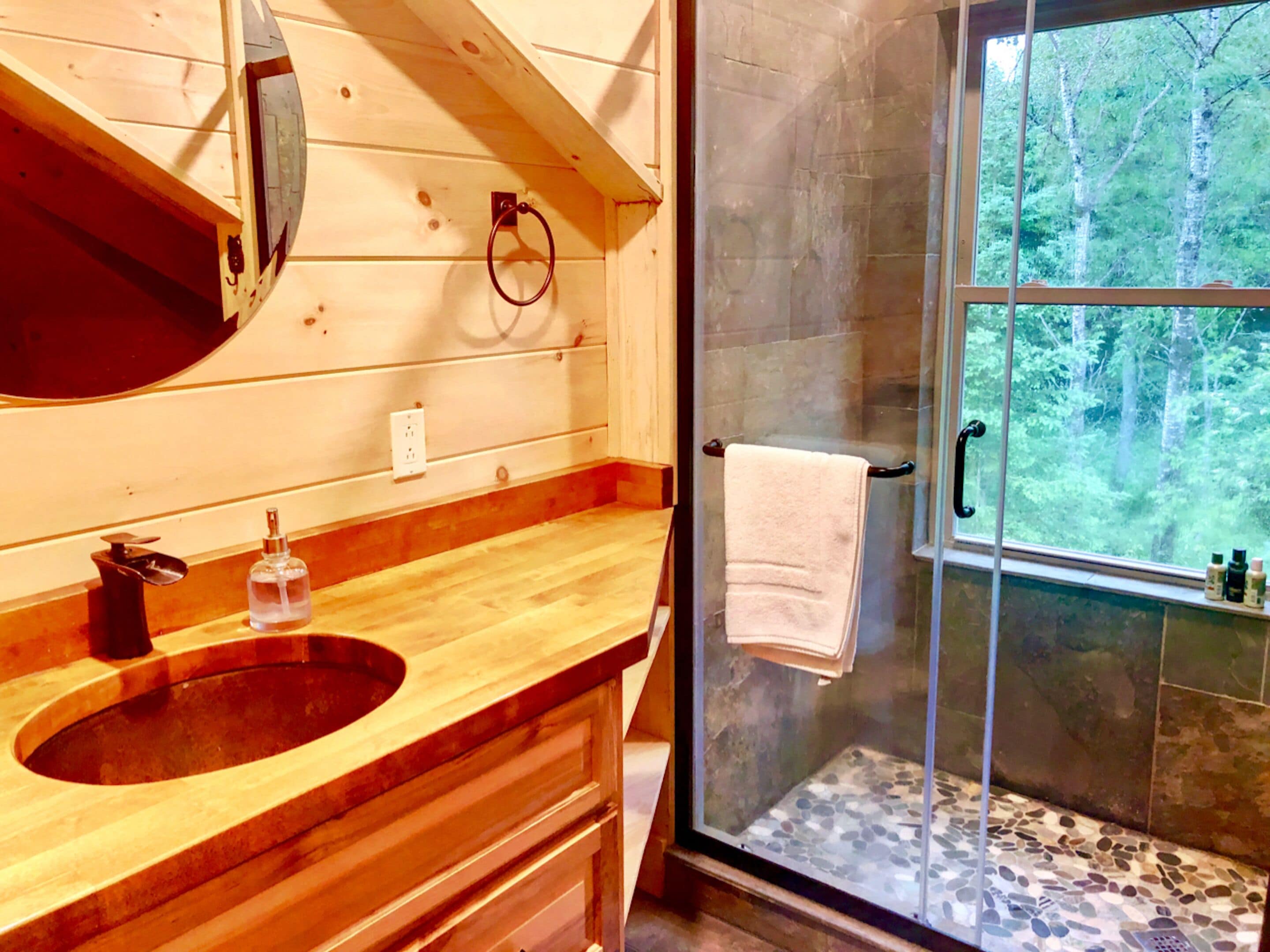 Une salle de bain en bois avec un grand lavabo, un miroir et une douche en verre donnant sur la forêt.