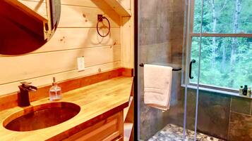 Une salle de bain en bois avec un grand lavabo, un miroir et une douche en verre donnant sur la forêt.