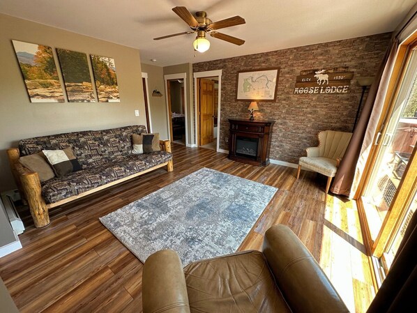 A cozy living room with a wooden floor, a patterned sofa, a fireplace, and a large window.
