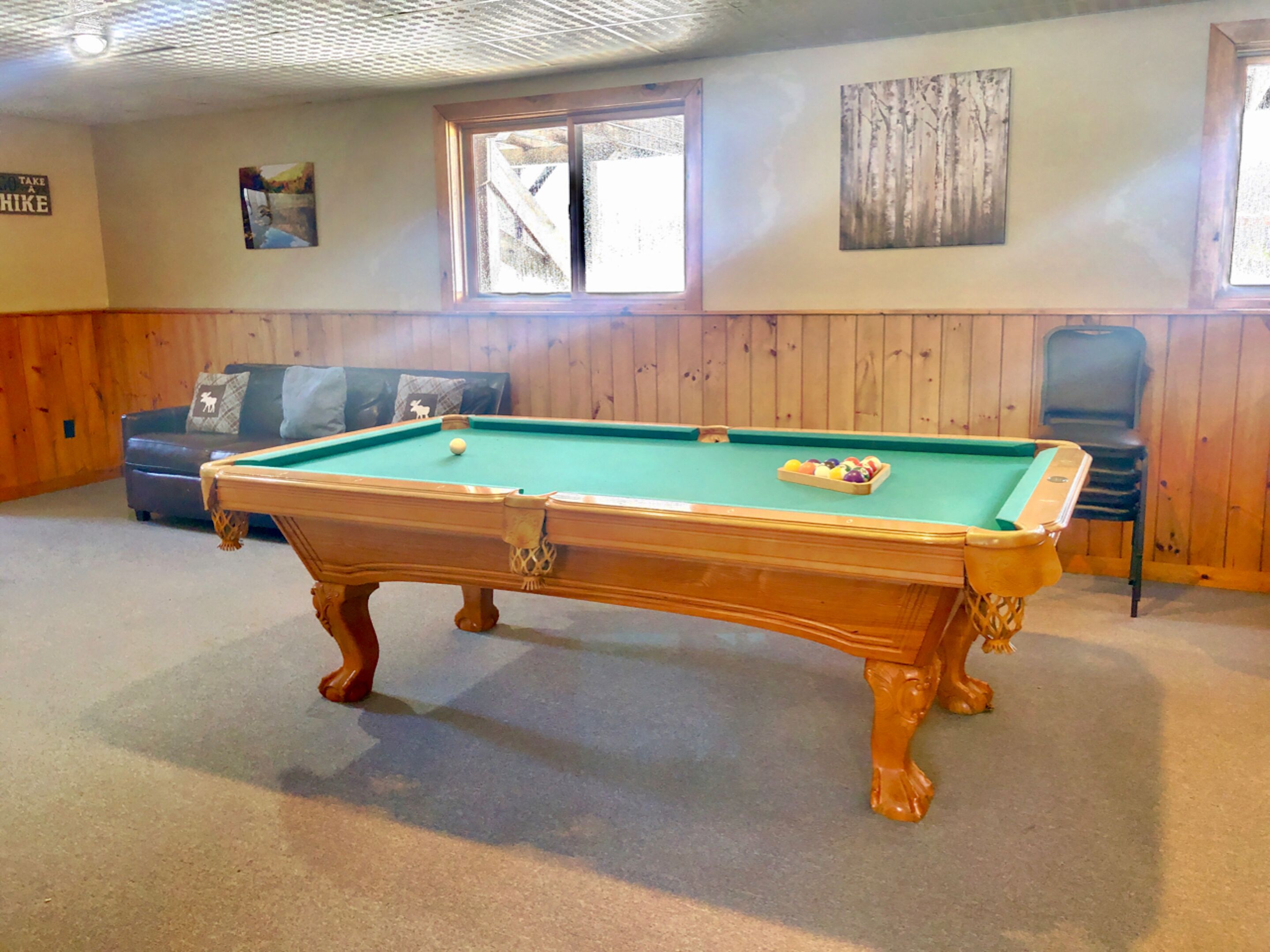 Una mesa de billar en una habitación con paneles de madera, un sofá y una ventana.