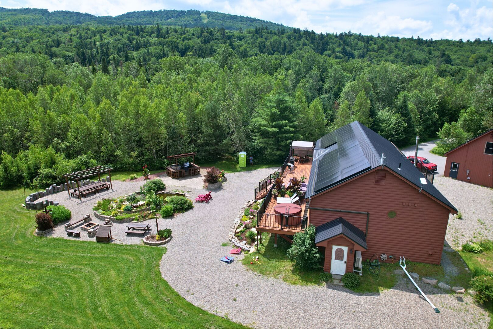 A rustic cabin with a deck, surrounded by a forested area and a gravel driveway.