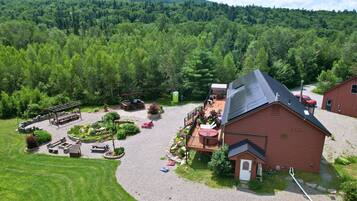 A rustic cabin with a deck, surrounded by a forested area and a gravel driveway.