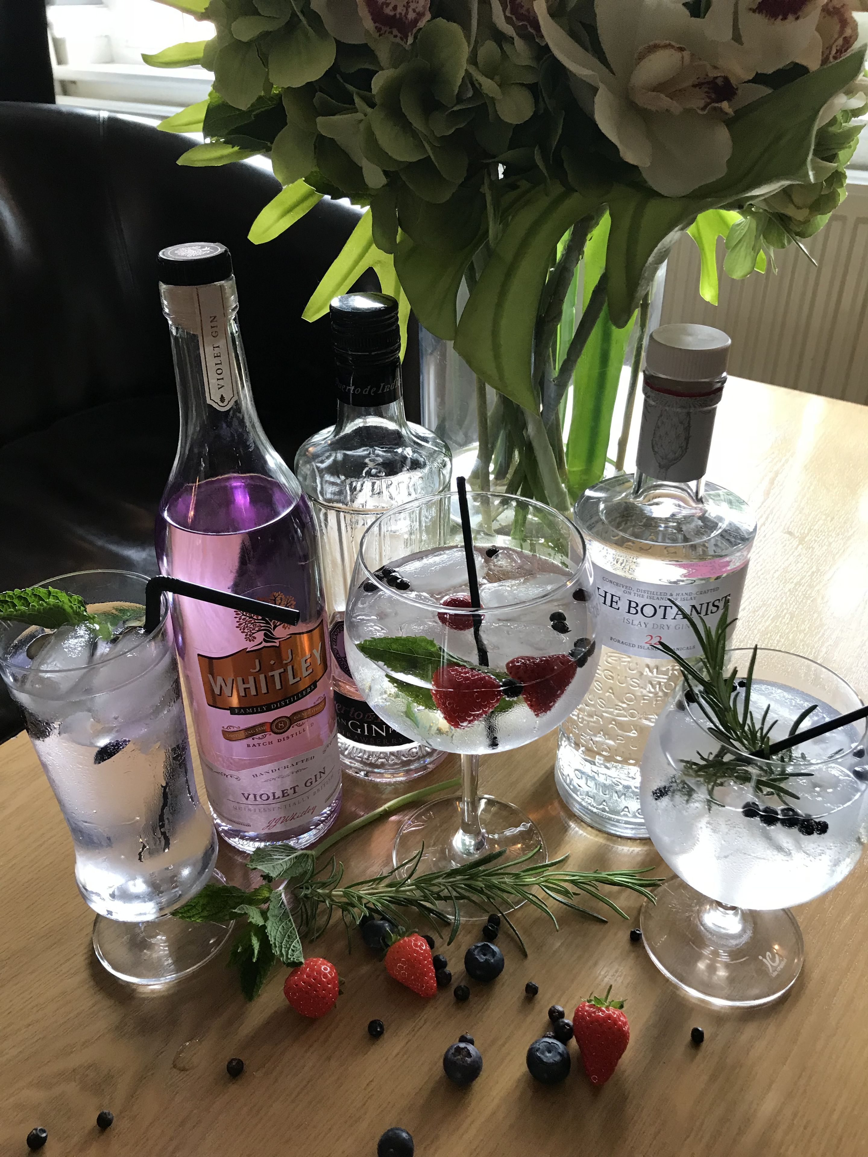 Una mesa con varias botellas de ginebra, dos vasos con hielo y bayas, y un florero con flores.