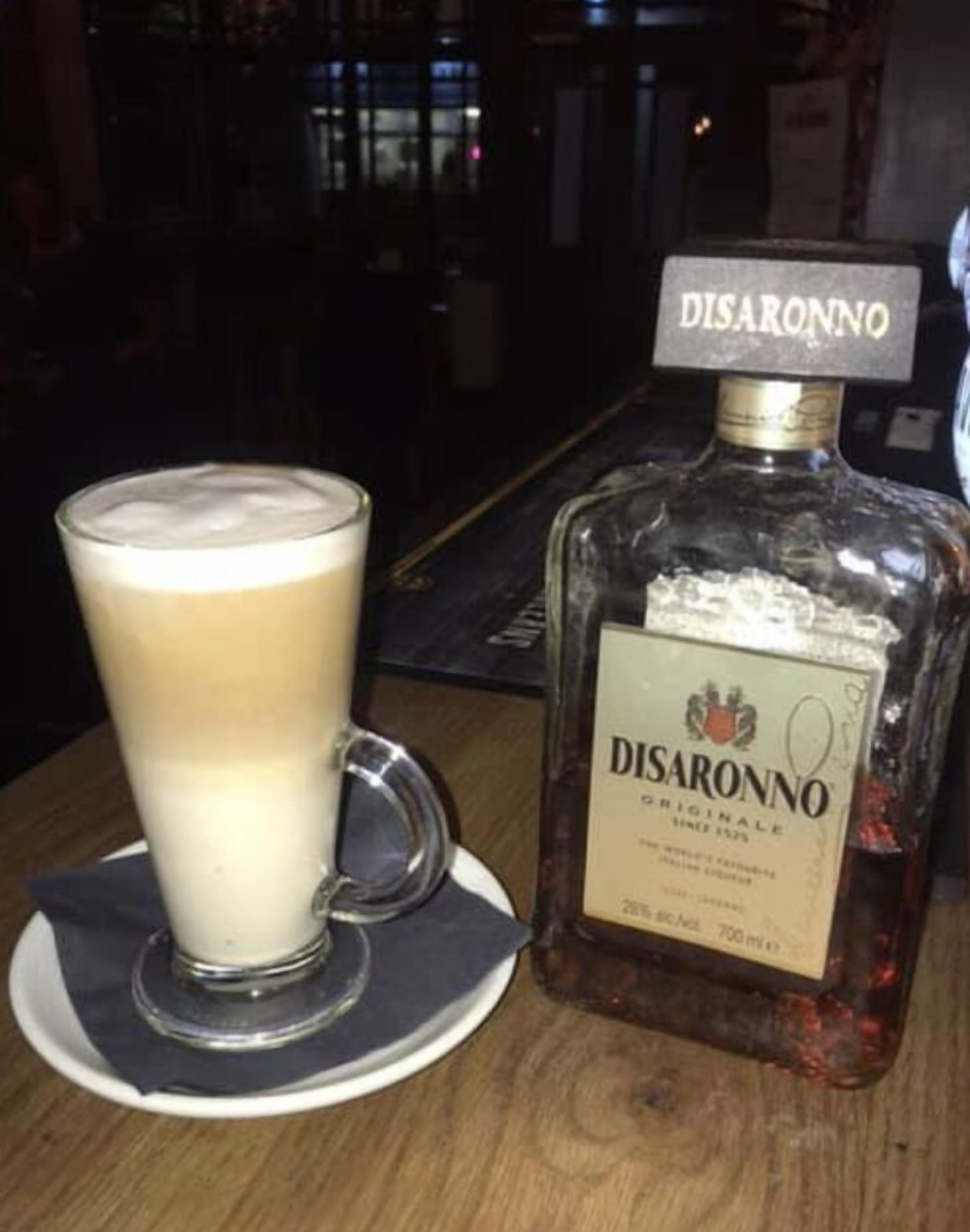 A glass of coffee with a foamy top, a bottle of Disaronno, and a coaster on a wooden table.