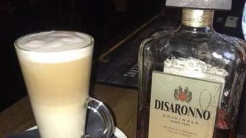 A glass of coffee with a foamy top, a bottle of Disaronno, and a coaster on a wooden table.