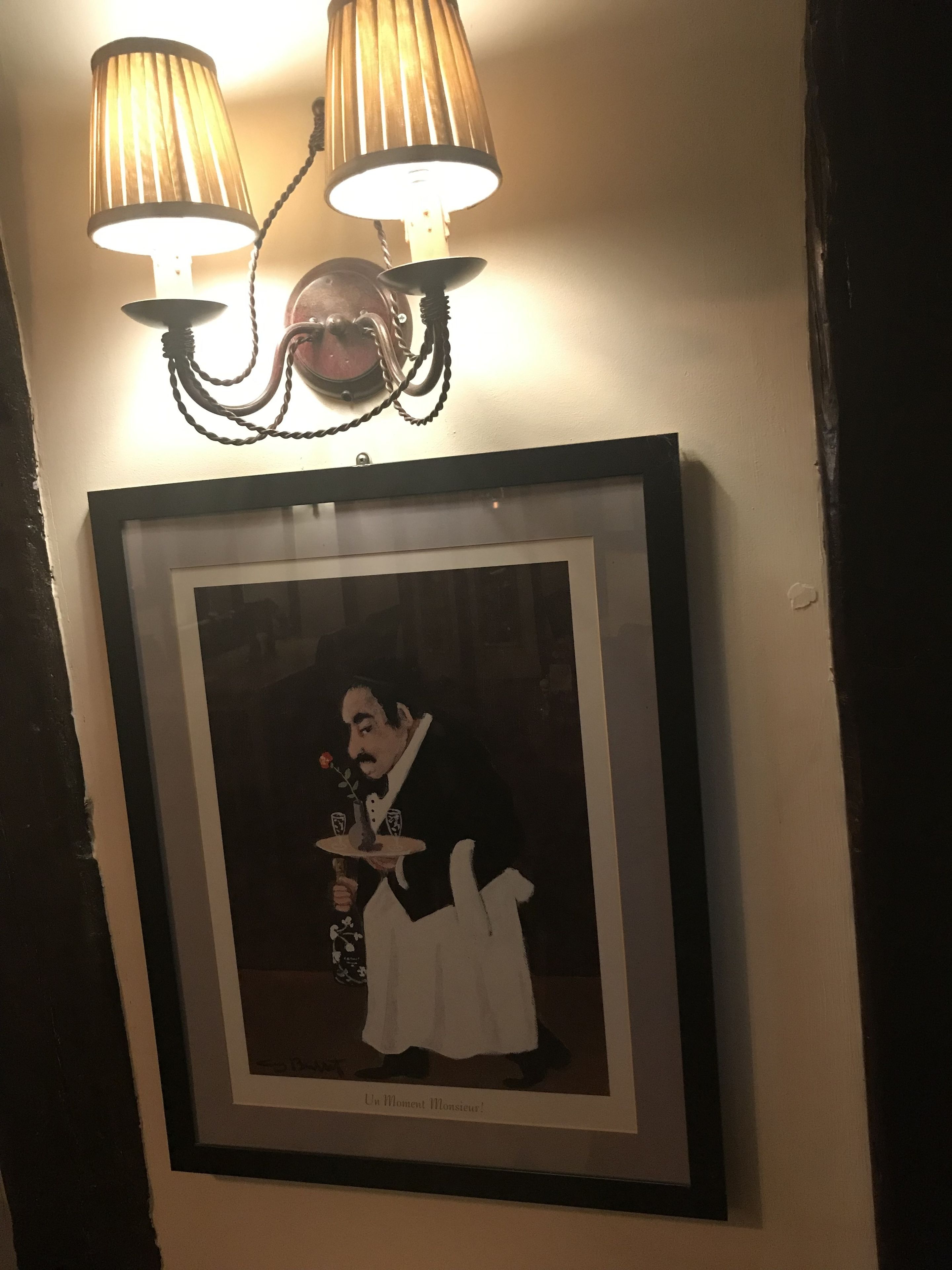 A framed artwork of a man in a black suit holding a tray with a rose, hanging on a wall above a dark wooden door.