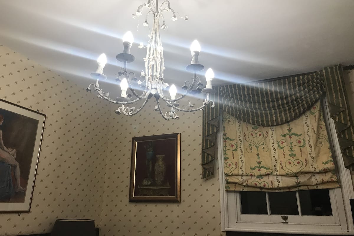 Quarto com lustre, papel de parede estampado, quadro emoldurado e janela com cortinas.