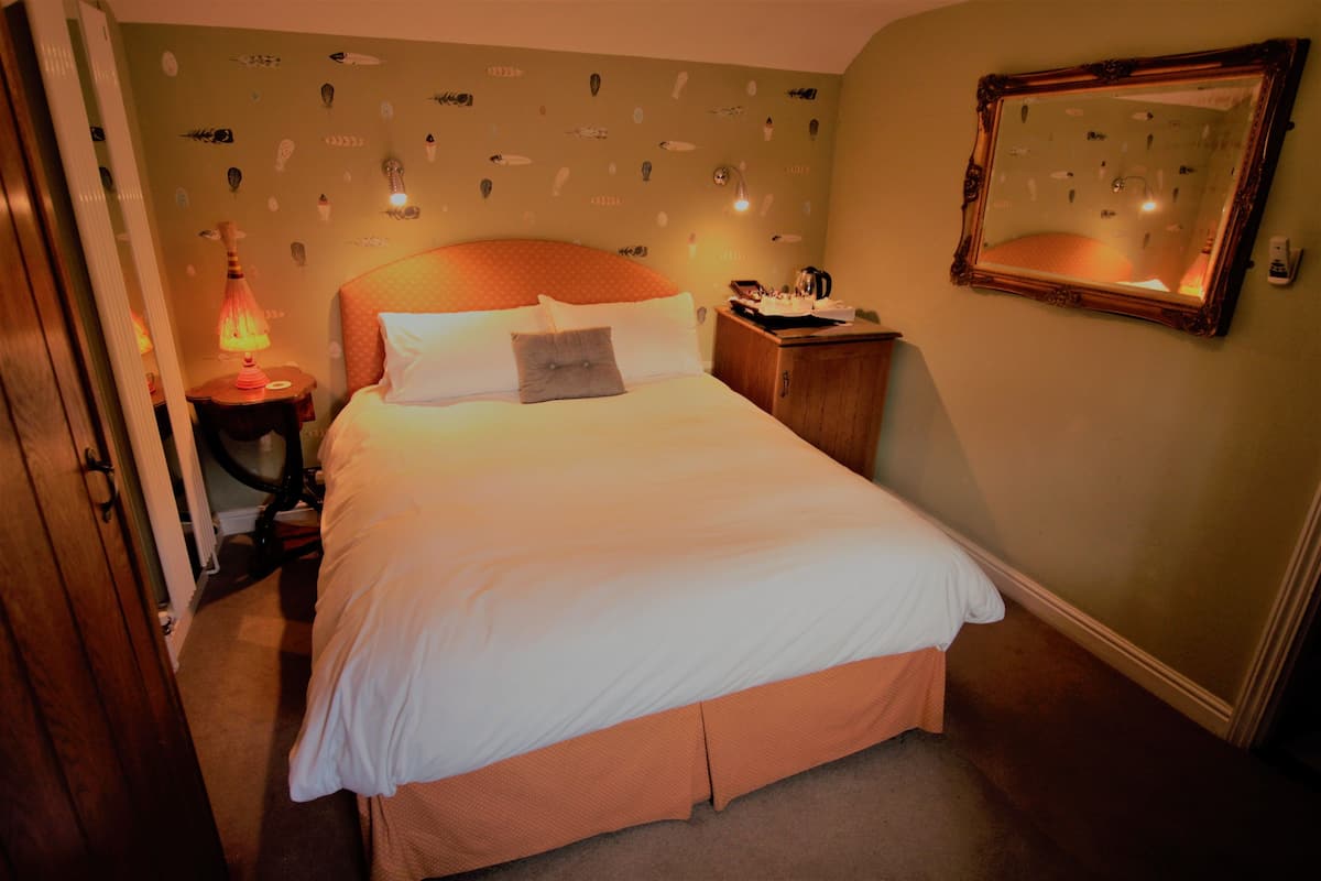 Uma cama bem arrumada com roupa de cama branca e cabeceira de madeira, uma mesa de cabeceira com abajur e um espelho fixado na parede.