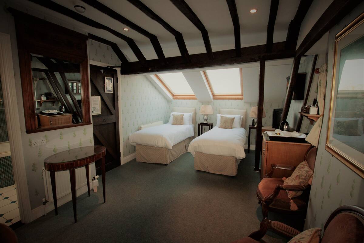 Quarto com duas camas, uma mesa, uma cadeira e um aparador. Há um claraboia sobre as camas e uma janela à esquerda.