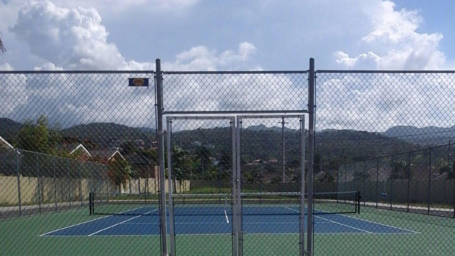 Un campo da tennis con una recinzione metallica e un cartello, circondato da case e montagne.