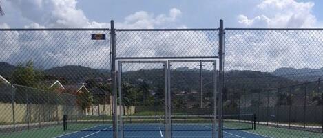 Un campo da tennis con una recinzione metallica e un cartello, circondato da case e montagne.