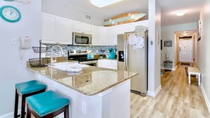 A modern kitchen with white cabinets, a granite countertop, and a built-in microwave. There are two teal bar stools and a small island with a sink. - Beachfront Gulf Shores Condo w/ Pool (Gulf Shores)