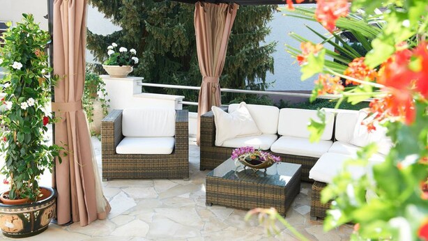 An outdoor seating area with wicker furniture, white cushions, and a stone floor. - Hotel Wendelstein (Wendelstein)